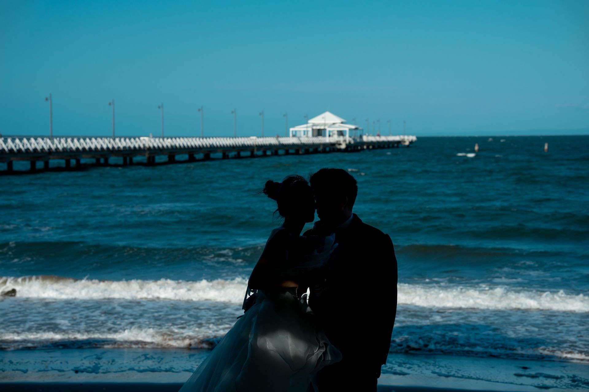 Silhouette of a couple embracing on a beach with a pier in the background.
