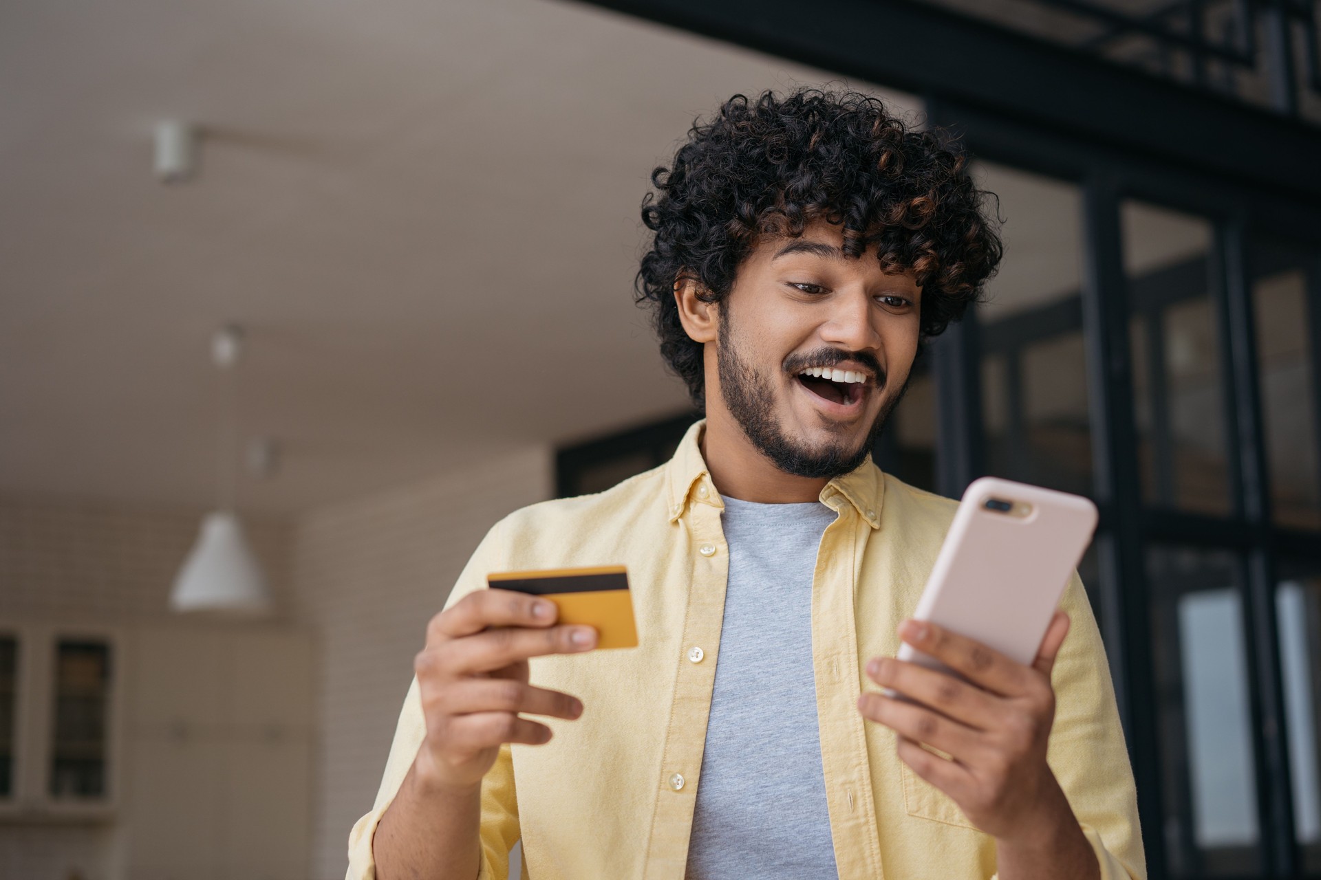 Indian man holding credit card using mobile phone shopping online. Happy freelancer receive payment, check card balance. Emotional guy ordering food online looking at digital screen